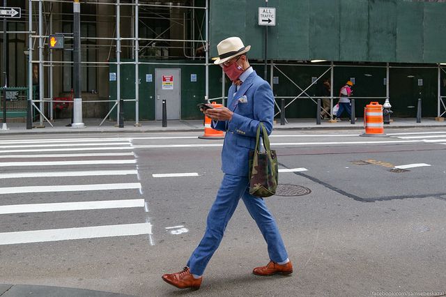 A well-dressed man wearing a suit and face mask walks down a city street while staring at his phone in his hands.